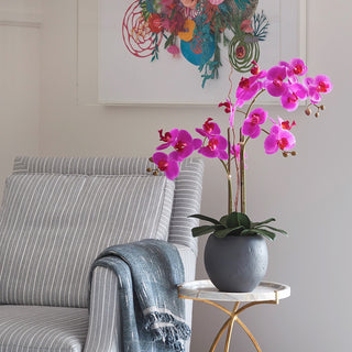 Beautiful setting featuring the Artificial Pink Double Stem Orchid by CG Hunter beside a cozy chair, adding a touch of luxury to a refined living space.