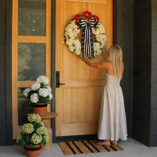 30-inch artificial white hydrangea wreath with navy ribbon hung on a front door beside potted hydrangea plants 30” Artificial White Hydrangea Wreath styled for the Fourth of July. Featuring a ribbon by Tableshoppeco.