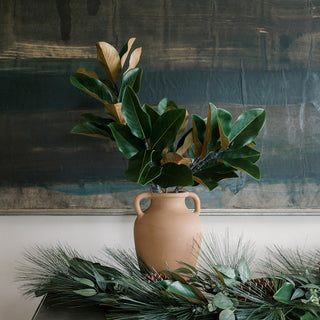 Elegant arrangement of magnolia leaves in a stylish vase atop a luxurious table, complemented by faux holiday greenery, showcasing pieces from the Christmas Greenery collection by CG Hunter.