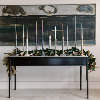 Elegant display of CG Hunter handcrafted candleholders arranged on a stylish black console table, featuring an exquisite assortment of taper candles in various colors, complemented by a modern art backdrop and lush greenery, showcasing luxury home décor.