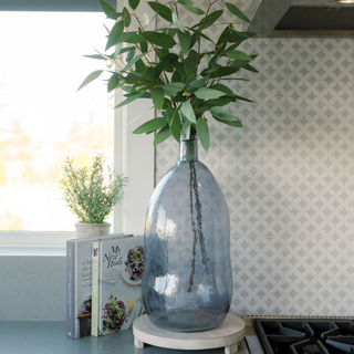 Sophisticated CG Hunter Hammered Glass Vase in Smoke on a wooden base in a contemporary kitchen, elegantly holding greenery and positioned beside decorative books.