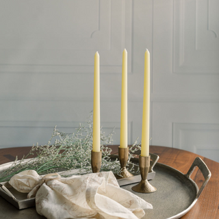 Luxurious arrangement featuring CG Hunter's 3.5" Iron Candleholders in Antique Brass holding soft yellow taper candles on a decorative tray surrounded by delicate greenery.