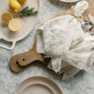 Luxurious CG Hunter Grey and Green Lilies Tablecloth and Napkins, elegantly arranged with fresh fruits on a wooden serving board, showcasing delicate floral designs for a sophisticated dining experience.