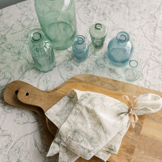 Stylish display of the CG Hunter Grey and Green Lilies Tablecloth and Napkins, set against an array of soft blue glass vases on a wooden cutting board, evoking a sophisticated dining aesthetic.