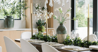 Dining table set with greenery and decorative items in a bright room.