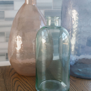 Artful arrangement of CG Hunter Handmade Hammered Glass Vases in Peach and other colors on a wooden table, showcasing artistic craftsmanship and adding charm to a modern interior.