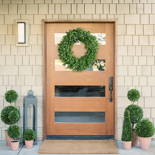Wooden front door with a wreath, flanked by potted plants against a beige tiled wall.