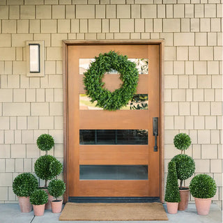 Wooden front door with a green wreath, flanked by potted plants on a beige tiled wall background.