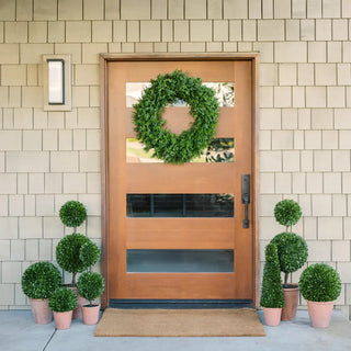 Wooden front door with a green wreath, surrounded by potted plants on a tiled wall background.
