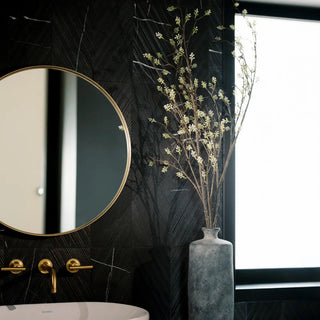 Round mirror with gold frame on a black wall next to a vase with flowers.