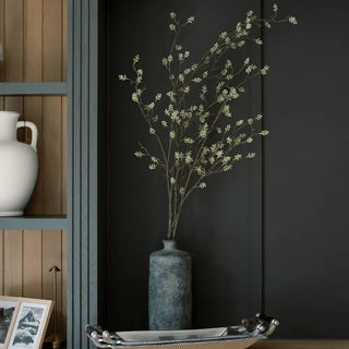 Decorative arrangement of branches in a vase against a dark wall with a shelf and decorative items.