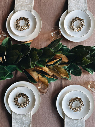 Four exquisite 7.5" Iron Wreath Ornaments in brass by CG Hunter, beautifully arranged on a dining table with fine china and lush green leaves, perfect for sophisticated holiday gatherings.