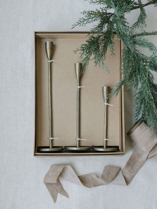 Set of three handcrafted antique brass candlesticks by CG Hunter, beautifully packaged in a box with a decorative green branch accent.