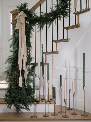 Stylish staircase adorned with handcrafted antique brass candlesticks by CG Hunter, displaying an elegant assortment of taper candles amidst seasonal garland.