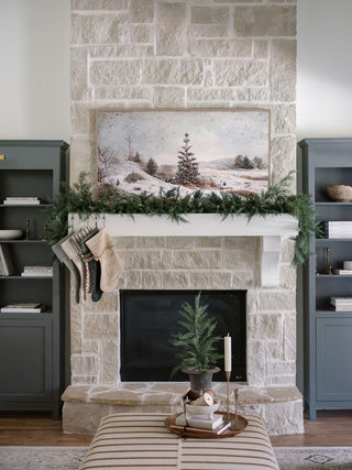 A refined living room showcasing a 9' Faux Cedar Garland by CG Hunter, elegantly draped over a stone fireplace, complemented by decorative stockings and modern decor, creating a warm festive atmosphere.