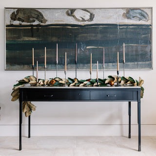 Elegant display featuring a 9' Faux Green Magnolia Leaf Garland by CG Hunter, draped across a stylish black table with candles, set against a modern artwork.