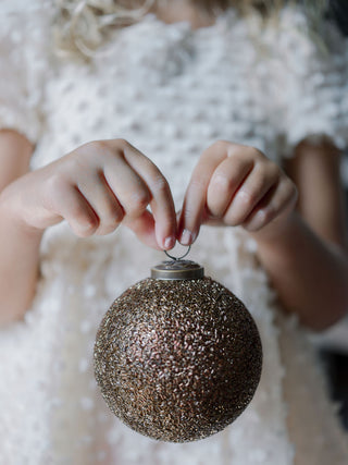 A child's delicate hands hold a beautiful 4" Beaded Antique Bronze Glass Ornament by CG Hunter, symbolizing the joy and luxury of festive decorating with handcrafted pieces.