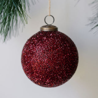 Close-up of a luxurious 4" Beaded Bordeaux Glass Ornament by CG Hunter, featuring a rich textured surface and an antique bronze topper, elegantly displayed against a backdrop of evergreen branches.