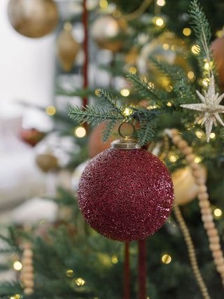 A stunning 4" Beaded Bordeaux Glass Ornament by CG Hunter hanging from a beautifully decorated Christmas tree, showcasing its shimmering texture and adorned with twinkling lights.