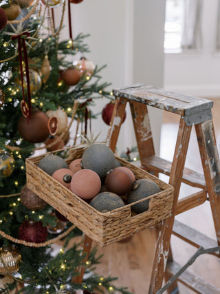 A woven basket filled with various luxurious ornaments, including the 5" Textured Slate Blue Glass Ornament by CG Hunter, set near a beautifully decorated Christmas tree.