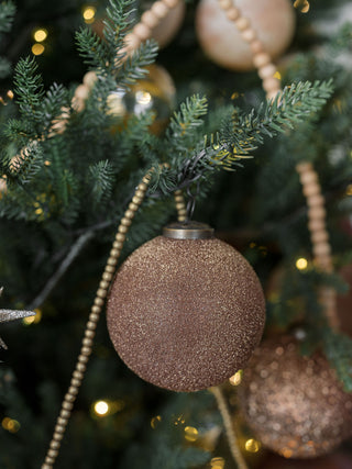 Close-up of a 5-inch textured cinnamon glass ornament by CG Hunter, featuring a shimmering surface, hanging elegantly on a decorated evergreen tree.