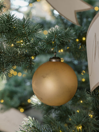 4" Matte Antique Gold Glass Ornament by CG Hunter, artfully displayed amidst a lush green Christmas tree, illuminated by soft lights, enhancing festive ambiance.