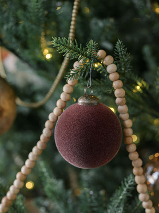 Stylish 3" Flocked Bordeaux Glass Ornament by CG Hunter, adorning a tree branch amid wooden beads, enhancing the festive atmosphere with its rich color and texture.