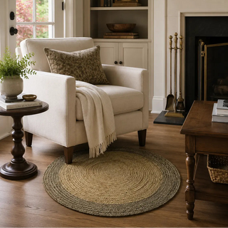 A cozy living room scene featuring a cream upholstered armchair with a patterned throw pillow and a cream fringed throw blanket, placed on a round braided jute rug. The room includes a wooden side table, a dark wood coffee table, a fireplace with gold-toned tools, and built-in shelves in the background.