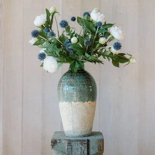 Decorative vase with green and beige base holding a bouquet of white and blue flowers against a neutral background.