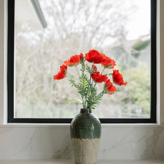 Green vase with red flowers on a marble surface in front of a window