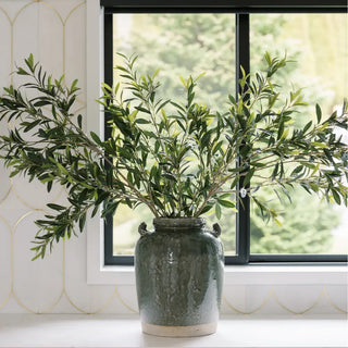 Green vase with olive branches on a windowsill with a blurred outdoor background