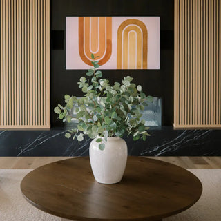 White vase with green plant on a wooden table against a black wall with abstract art