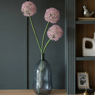 Clear glass vase with pink flowers against a dark gray wall with a shelf in the background.