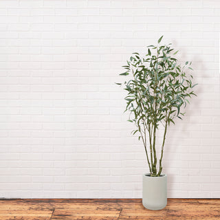 Minimalist decor showcasing the 7' Artificial Willow Eucalyptus Tree with Artisan Planter by CG Hunter, enhancing a modern space against a white brick wall.