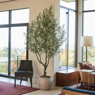 Stunning living area featuring CG Hunter Artificial Olive Tree 8.5' in Artisan Mediterranean Pot, complementing a modern lounge chair and large windows with scenic views.