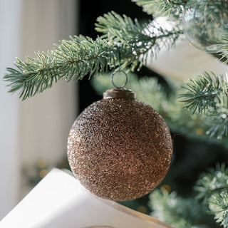 Close-up of a stunning 4" Beaded Antique Bronze Glass Ornament by CG Hunter, elegantly hanging on a lush green tree branch, highlighting its rich texture and antique finish.