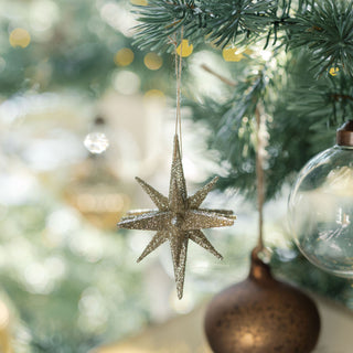 A sparkling Gold Glitter Starburst Ornament gracefully dangling from a Christmas tree branch, surrounded by decorative bulbs and soft lighting, emphasizing the elegance of CG Hunter's festive collection.