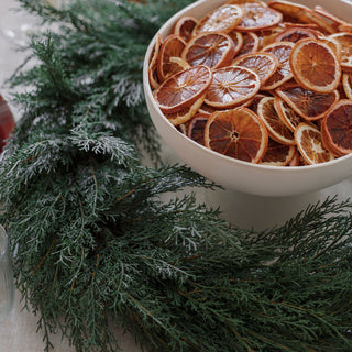 A luxurious 30" Faux Evergreen Cedar Wreath from CG Hunter beautifully enhances a festive table setting with a bowl of dried orange slices, showcasing the wreath's lifelike greenery.
