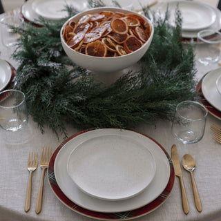 A sophisticated dining arrangement featuring a 30" Faux Evergreen Cedar Wreath from CG Hunter as a centerpiece, surrounded by plates and gold utensils, creating a warm holiday ambiance.