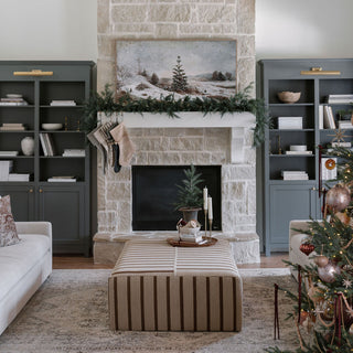 An elegant living room featuring a stylish stone fireplace adorned with a stunning artwork of a snowy cedar tree scene. The cozy decor includes a beautiful arrangement of lifelike cedar trees and a luxurious cedar wreath from CG Hunter, enhancing the natural beauty and sophistication of the space. The tasteful furnishings and festive touches create a warm, inviting atmosphere perfect for holiday gatherings.