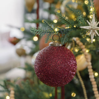 A stylish look at the 4" Beaded Bordeaux Glass Ornament by CG Hunter, gracefully suspended among holiday greenery and sparkling lights, emphasizing its exquisite craftsmanship.