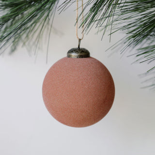 A luxurious 3" Flocked Vintage Rose Glass Ornament by CG Hunter, showcasing a soft rose hue and velvety texture, hanging delicately from a pine branch, perfect for enhancing holiday decor.