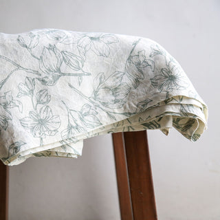 Close-up of the CG Hunter Grey and Green Lilies Tablecloth draped gracefully over a wooden stool, showcasing the intricate lily pattern and luxurious linen texture.