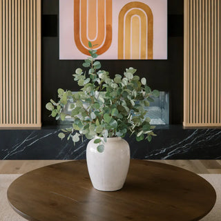 White vase with green plant on a wooden table against a black wall with abstract art