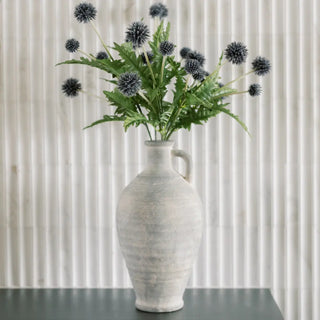 White textured vase with green leaves and black thistle-like flowers against a neutral background