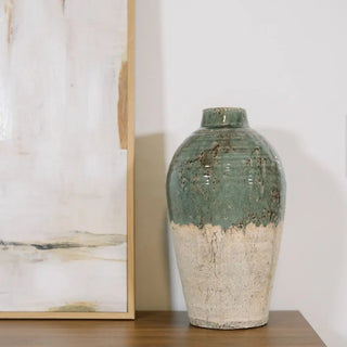 Green ceramic vase on a wooden surface with a blurred abstract painting in the background