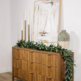 Wooden dresser with decorative greenery, candles, and a vase against a white wall.