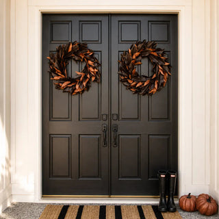 A pair of black double front doors with decorative leaf wreaths hanging on each door, set in a white entryway. A striped natural fiber doormat, black rain boots, and small pumpkins sit on the porch floor.
