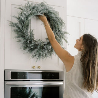 Stylish 30" Flocked Cedar Wreath by CG Hunter being positioned above cabinetry, enhancing the decor with its frosted finish and realistic detail.