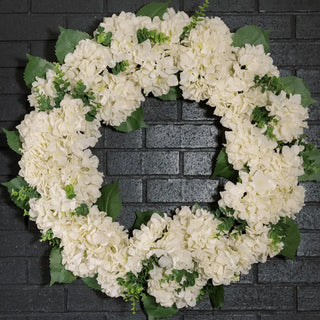 Floral wreath made of white flowers and green leaves against a dark brick wall.